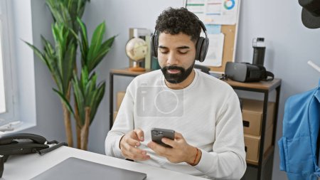 Téléchargez les photos : Un jeune hispanique barbu utilise un smartphone dans un bureau moderne, entouré d'accessoires technologiques et de plantes. - en image libre de droit
