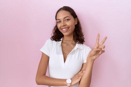 Téléchargez les photos : Jeune femme hispanique portant un t-shirt blanc décontracté souriant avec un visage heureux clin d'oeil à la caméra faisant signe de victoire avec les doigts. numéro deux. - en image libre de droit