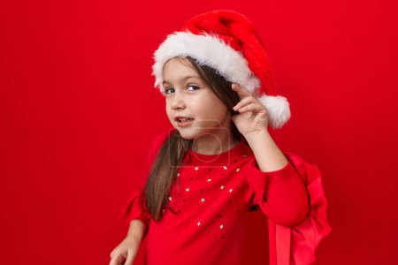 Foto de Linda chica hispana en el sombrero de Navidad sobre-escuchar chismes, de la mano en la oreja, sordera insinuó. cautivante escena sobre rojo aislado fondo - Imagen libre de derechos