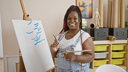 Photo for Joyful african american female artist draws confidently, smiling radiantly in her art studio while standing over her canvas, embracing her creativity - Royalty Free Image