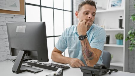 Photo for Pensive tattooed hispanic man in a modern office, contemplating while sitting at his desk with a computer. - Royalty Free Image