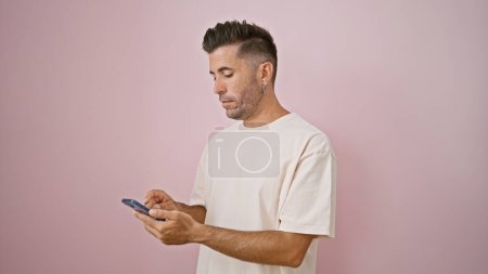 Foto de Un joven hispano de cara seria, absorto en una intensa conversación digital por teléfono. sosteniendo su teléfono inteligente, está escribiendo un mensaje, aislado contra una pared de fondo rosa. - Imagen libre de derechos
