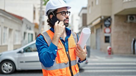 Photo for Young hispanic man architect holding blueprints talking on smartphone at construction place - Royalty Free Image