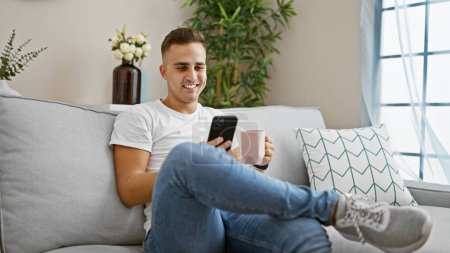 Photo for Young hispanic man smiling using smartphone, relaxing on sofa at home with coffee mug. - Royalty Free Image