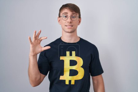 Téléchargez les photos : Homme blond caucasien portant Bitcoin t-shirt sourire et geste confiant avec la main faisant signe de petite taille avec les doigts à la recherche et la caméra. concept de mesure. - en image libre de droit
