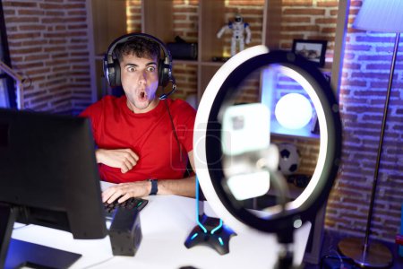 Photo for Young hispanic man playing video games recording with smartphone scared and amazed with open mouth for surprise, disbelief face - Royalty Free Image