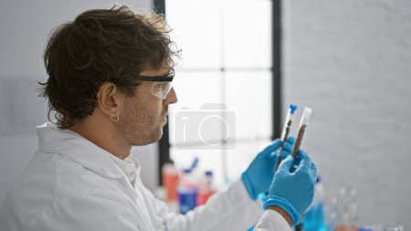 Foto de Un hombre barbudo con gafas de seguridad y guantes examina los tubos de ensayo en un entorno de laboratorio brillante. - Imagen libre de derechos
