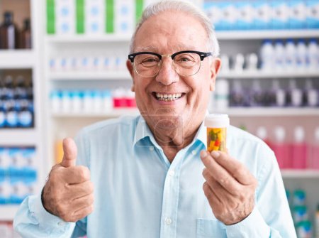 Foto de Hombre mayor con pelo gris sosteniendo pastillas en la farmacia sonriendo feliz y positivo, pulgar hacia arriba haciendo excelente y signo de aprobación - Imagen libre de derechos