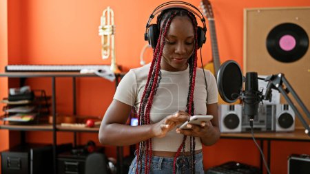 Téléchargez les photos : Mélodie sincère, musicienne afro-américaine chantant avec âme, regardant l'écran du smartphone dans un studio de musique - en image libre de droit