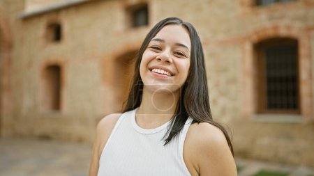 Foto de Sonriente joven hispana llena de alegría se encuentra confiada en una calle urbana, su expresión feliz capturando su estilo de vida fresco e informal. este hermoso retrato muestra su felicidad al aire libre. - Imagen libre de derechos