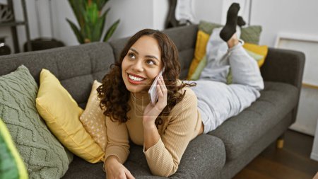 Photo for Smiling woman talking on phone while relaxing on a sofa in a cozy living room, portraying a casual, comfortable lifestyle. - Royalty Free Image