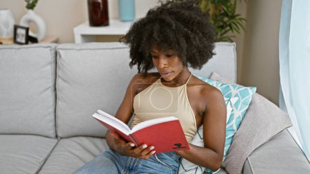 Foto de African american woman reading book sitting on sofa at home - Imagen libre de derechos