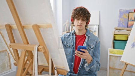 Téléchargez les photos : Un jeune homme aux cheveux bouclés portant des lunettes et une veste en denim vérifiant son smartphone dans un studio d'art créatif - en image libre de droit