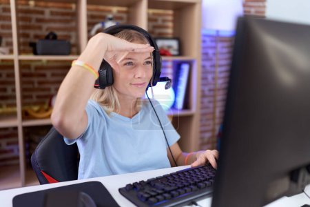 Téléchargez les photos : Jeune femme caucasienne jouant à des jeux vidéo portant des écouteurs très heureux et souriant regardant loin avec la main sur la tête. concept de recherche. - en image libre de droit