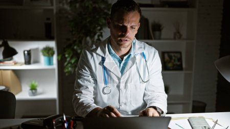 Foto de Un hombre hispano enfocado en una bata de laboratorio trabaja hasta altas horas de la noche en una clínica, iluminado por el resplandor de una pantalla de computadora. - Imagen libre de derechos