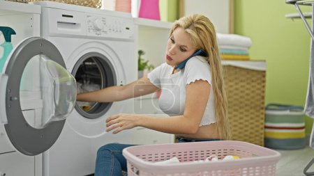 Photo for Young blonde woman talking on smartphone washing clothes at laundry room - Royalty Free Image