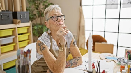 Foto de Intrigante escena en la escuela de arte, anciana artista de cabello gris sentada pensativamente en una mesa, dudando de su siguiente pincelada en clase de arte - Imagen libre de derechos