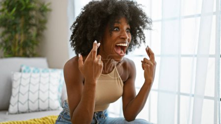 Téléchargez les photos : Femme afro-américaine souriante confiante assise sur un canapé croisant les doigts à la maison - en image libre de droit