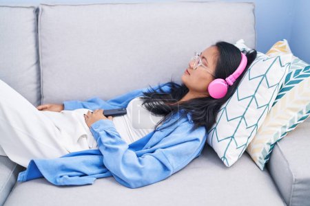 Téléchargez les photos : Jeune femme chinoise écoutant de la musique dormir sur le canapé à la maison - en image libre de droit