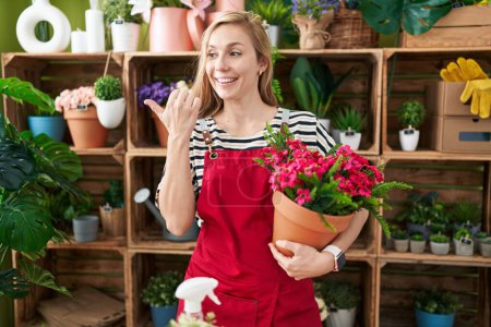Foto de Joven mujer caucásica trabajando en floristería sosteniendo planta apuntando pulgar hacia el lado sonriendo feliz con la boca abierta - Imagen libre de derechos