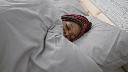 Foto de Mujer afroamericana agotada en trenzas, cómodamente acostada en una cama acogedora, durmiendo en su cálido y relajante dormitorio, rindiéndose a la dulce llamada del sueño después de un día agotador. - Imagen libre de derechos