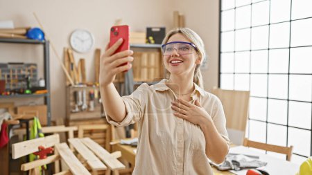Téléchargez les photos : Femme souriante prend un selfie dans un atelier avec des outils de menuiserie sur un établi derrière elle. - en image libre de droit