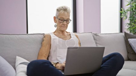 Photo for Focused elderly woman with grey hair and glasses, using laptop with concentration while sitting on sofa indoors at home, embodying mature technology use in the living room - Royalty Free Image
