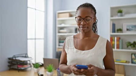 Téléchargez les photos : Femme afro-américaine avec des lunettes utilisant un smartphone dans un cadre de bureau moderne. - en image libre de droit