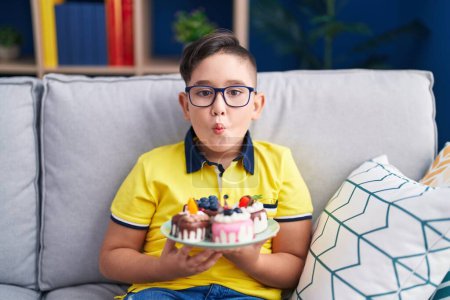 Téléchargez les photos : Jeune enfant hispanique tenant bonbons gâteau faisant visage de poisson avec la bouche et les yeux plissés, fou et comique. - en image libre de droit