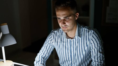 Téléchargez les photos : Jeune homme concentré travaillant tard dans un bureau moderne, éclairé par un écran et une lampe de bureau. - en image libre de droit