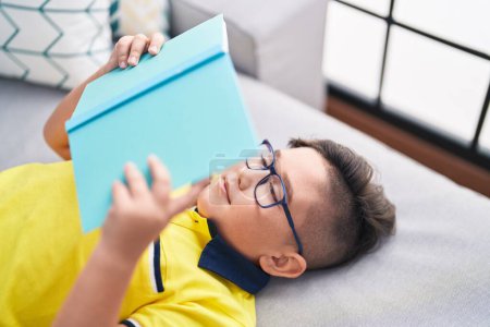 Photo for Adorable hispanic boy reading book lying on sofa at home - Royalty Free Image