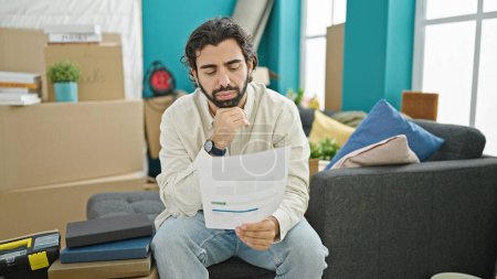 Téléchargez les photos : Jeune homme hispanique lisant le document assis sur le canapé à la nouvelle maison - en image libre de droit