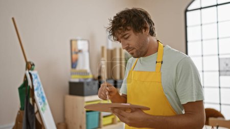 Photo for Focused man painting on canvas in a bright art studio wearing yellow apron. - Royalty Free Image
