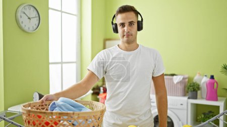 Photo for A young man in headphones holding a laundry basket stands in a brightly lit home laundry room. - Royalty Free Image