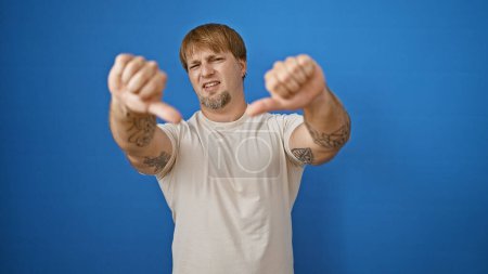 Bearded man with tattoos gives thumbs down against a blue background outdoors, expressing disapproval.