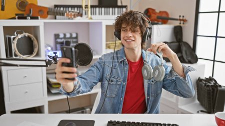 Foto de Un joven con auriculares se toma una selfie mientras disfruta de la música en un moderno estudio equipado con instrumentos. - Imagen libre de derechos