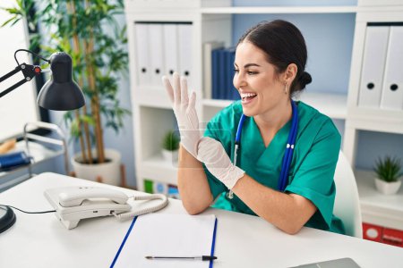 Téléchargez les photos : Jeune belle femme hispanique médecin souriant porter des gants confiants à la clinique - en image libre de droit