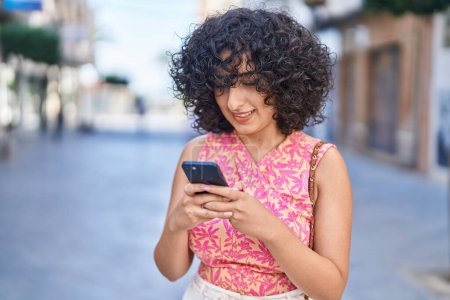 Téléchargez les photos : Jeune femme du Moyen-Orient souriant confiant en utilisant smartphone à la rue - en image libre de droit
