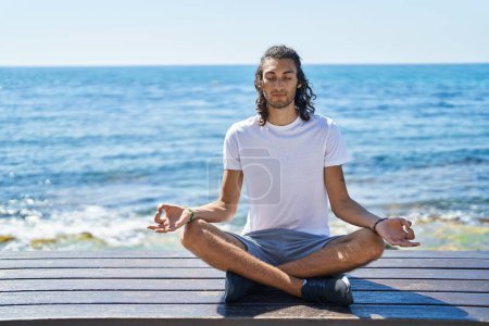Téléchargez les photos : Jeune homme hispanique faisant de l'exercice de yoga assis sur un banc au bord de la mer - en image libre de droit