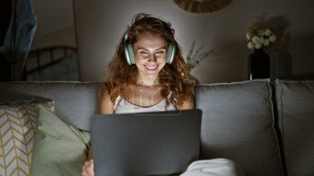 Photo for A smiling young woman enjoys music on headphones while using a laptop on the couch in a cozy living room interior. - Royalty Free Image