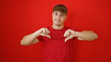 Photo for Cool young hispanic man making a negative thumbs down gesture, standing solo against a vivid red background. he embodies a casual fashion sense, expressing bad vibes or failure. - Royalty Free Image