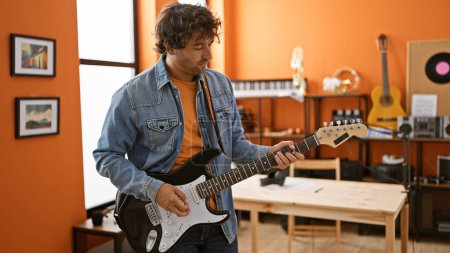 Téléchargez les photos : Un jeune hispanique joue de la guitare dans un studio de musique avec un design d'intérieur moderne et un équipement musical autour de lui. - en image libre de droit