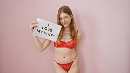 Foto de Mujer caucásica segura en lencería sosteniendo el signo de amor propio contra el fondo rosa - Imagen libre de derechos