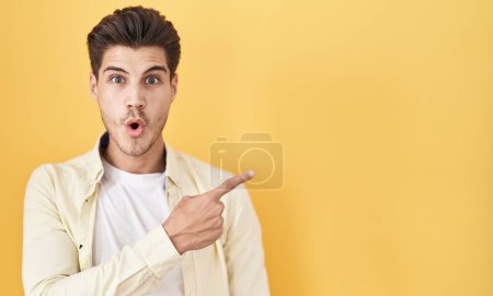 Foto de Joven hombre hispano de pie sobre fondo amarillo sorprendido señalando con el dedo hacia un lado, boca abierta expresión asombrada. - Imagen libre de derechos