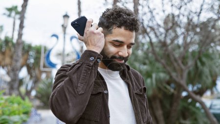Foto de Un apuesto joven hispano con barba disfrutando de una llamada telefónica al aire libre en un exuberante parque verde. - Imagen libre de derechos