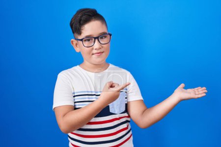 Téléchargez les photos : Jeune hispanique enfant debout sur fond bleu étonné et souriant à la caméra tout en présentant avec la main et pointant du doigt. - en image libre de droit
