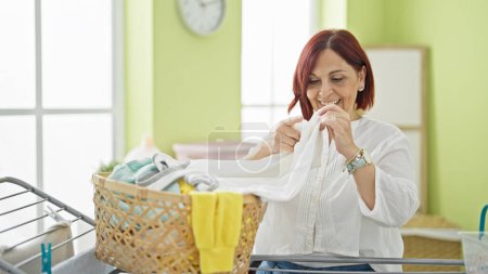 Foto de Mujer de mediana edad sonriendo confiada sosteniendo ropa de canasta en la lavandería - Imagen libre de derechos