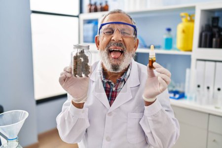 Foto de Hombre mayor hilarante divirtiéndose en el laboratorio, felizmente sacando la lengua con expresión divertida mientras sostiene la medicina del cannabis. - Imagen libre de derechos