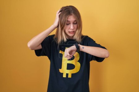 Foto de Mujer rubia caucásica vistiendo camiseta bitcoin mirando el tiempo reloj preocupado, miedo de llegar tarde - Imagen libre de derechos