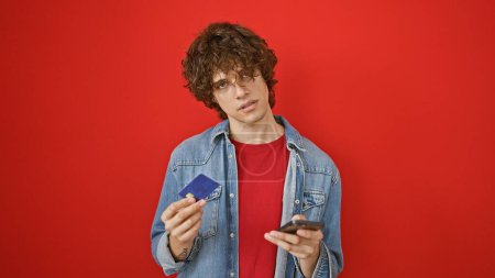 Téléchargez les photos : Jeune homme confus aux cheveux bouclés tenant une carte de crédit et un smartphone sur fond rouge - en image libre de droit